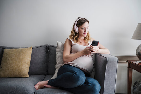 Pregnant woman listening music over headphones while using mobile phone on sofa at home