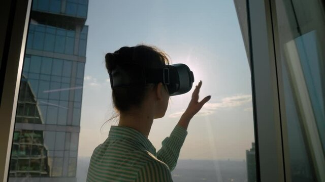 VR, opportunity, sightseeing, discover, technology concept. Woman using virtual reality headset and looking around against view on cityscape from panoramic skyscraper window in office. Sun lens flare