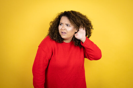 Young African American Woman Wearing Red Sweater Over Yellow Background Surprised With Hand Over Ear Listening An Hearing To Rumor Or Gossip