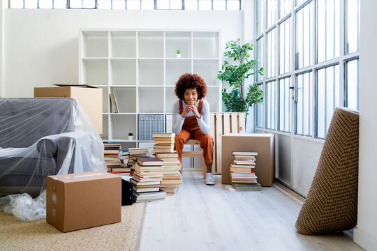 Smiling Afro woman with hand on chin sitting against bookshelf in new home