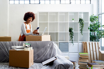 Smiling young woman standing with smart phone and laptop while moving into new home