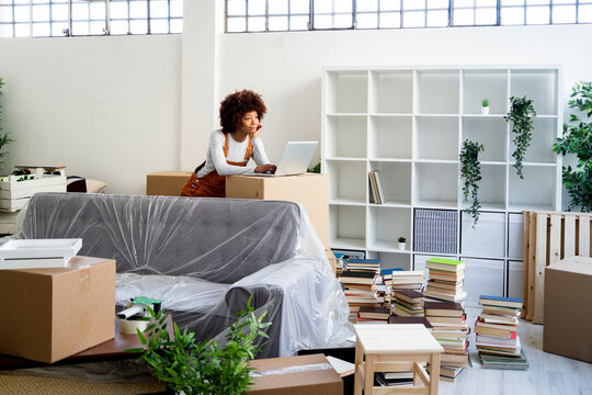 Afro woman with laptop leaning on cardboard box while moving into new home