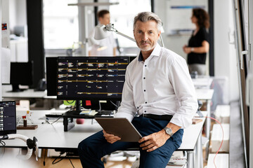 Male entrepreneur with digital tablet sitting on desk with colleagues in background at open plan office