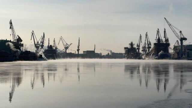 Cranes of of the Baltic shipyard on a frosty winter day, steam over the Neva river, smooth surface of the river, mirror reflection on the water, ships under construction, trawlers, nuclear icebreakers