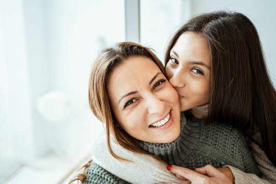 Daughter kissing happy mother by window at home