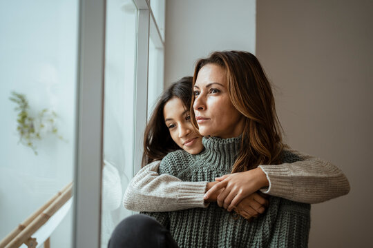 Daughter embracing mother while contemplating by window at home