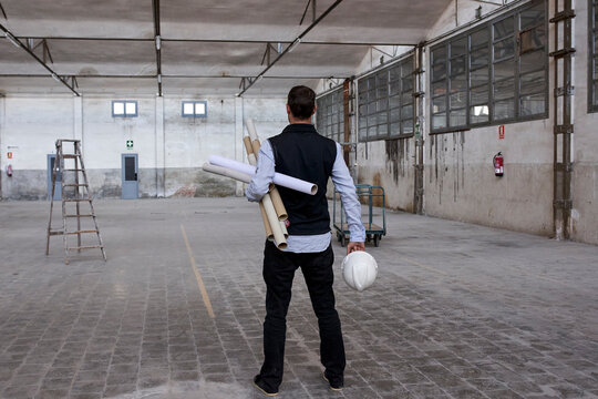 Male architect holding cardboards and hardhat while standing on floor in building