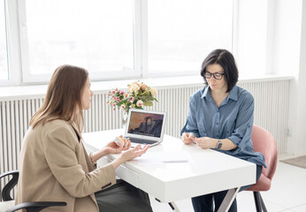 two young women conduct a coaching session in home office in light interior with laptop and flowers on tablework with metaphorical cards and psychological counseling