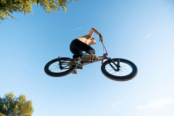 Caucasian white boy jumping with long hair shirtless with BMX bike on a mountain in the field with sand and trees blue sky sunny day