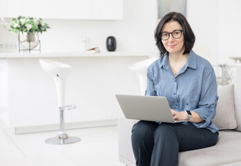 a young woman in glasses and in a blue shirt  works at home at a laptop as a coach in front of the kitchen in a bright interior, a concept of a workplace and remote work at home