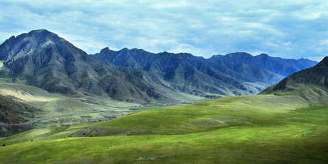 Alpine ridge with the Chuya river valley in Altai, in front of huge green hills, mountain road, sky with clouds, wide view, summer, cloudy