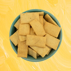 Cheese and garlic snack crackers in a green bowl on a yellow painted wood background top view.