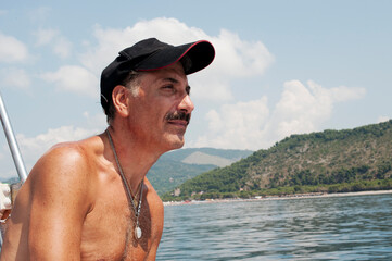 A Caucasian mustached man sitting on a motorboat's side enjoying the sea breeze and the panorama....
