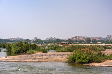 Anegundi, Karnataka, India - November 9, 2013: Sooryanarayana Temple island on gray Tungabhadra river under light blue sky. Green weeds on brown rocks.