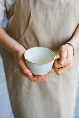 person in the apron holding handmade clay and turquoise cup of tea