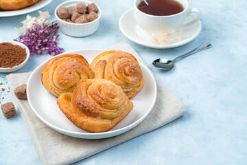 Three snail-shaped buns on a white plate against a background of a cup of tea and flowers. Side view with copy space.