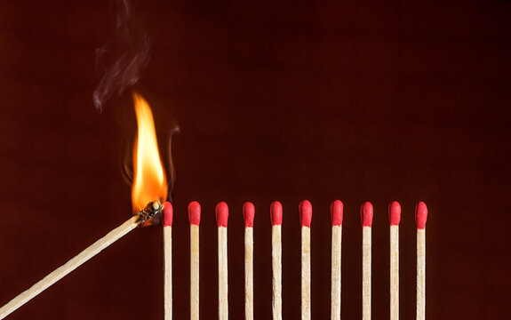 Lit match next to a row of unlit matches. Red phosphorus matches on dark red background. Concept of ignition or initiation