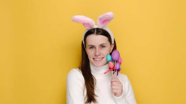 Good looking cute woman with pink bunny fluffy ears, has happy look, holds painted eggs on sticks, isolated on yellow studio background with copy space on right for your text. Holiday concept