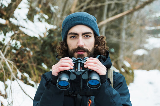 Male hiker in warm clothes standing in snowy winter woods and looking at camera holding binoculars