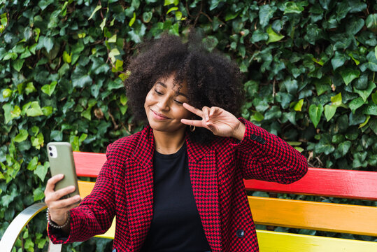 Content African American female sitting on bench in city and taking self portrait on smartphone while showing two fingers gesture