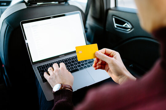 High angle of crop anonymous male entrepreneur sitting on backseat of luxury automobile with laptop and making online purchase while using plastic card