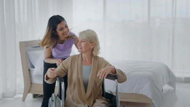 Joyful adopted Asian teen daughter comforts her injured Caucasian stepmother sitting in wheelchair by embracing and kissing her on the cheek. Love and caring time of Multi-racial family concept.