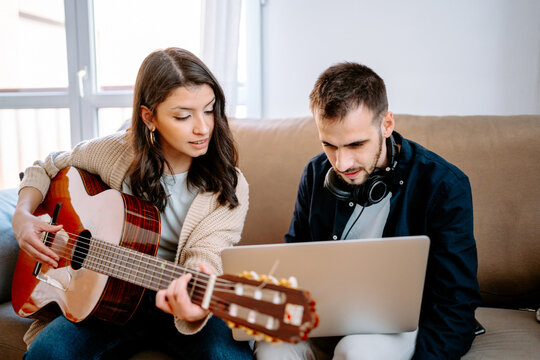 Couple of musicians sitting on sofa and recording song while playing acoustic guitar and using laptop
