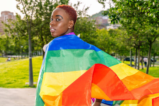 Side view of African American lesbian female wrapped in colorful LGBT flag standing on street and looking at camera