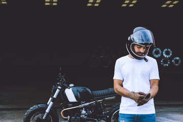 Full body of serious young ethnic male biker in casual clothes and cap standing near motorcycle near industrial cranes at seaside