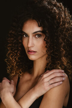 Serene seductive female with curly hair and brown eyes looking at camera on black background in dark studio