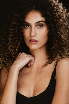 Serene seductive female with curly hair and brown eyes looking at camera on black background in dark studio