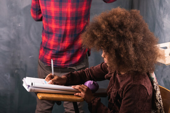 Cropped unrecognizable teacher writing mathematical example on chalkboard for black girl sitting at desk in classroom during lesson at school