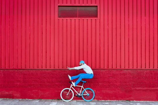 Side view of active bearded male cyclist in stylish outfit playing near bike on street near red building wall