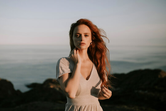 Beautiful young redhead unemotional woman on summer dress standing relaxing at mountain seaside landscape looking at camera with waving hair on background of blue sea