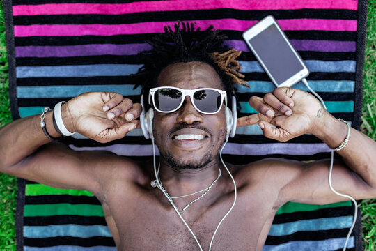 From above of carefree African American male lying on towel on lawn and enjoying songs in headphones in summer