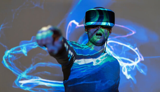 Unrecognizable young male in casual wear and VR headset getting new experience and touching virtual object in room with colorful projector illumination