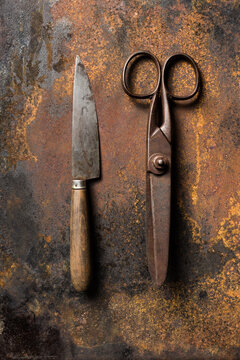 Top view of old metal scissors and sharp old knife placed on dark table with rusty surface