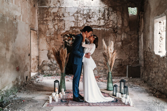Side view full body loving young ethnic newlywed couple wearing chic wedding clothes embracing and kissing gently in obsolete remaining building