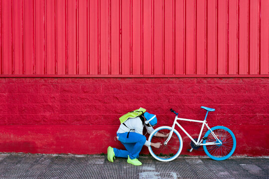 Side view of unrecognizable hipster man fixing wheel of modern bike on colorful red wall on the street