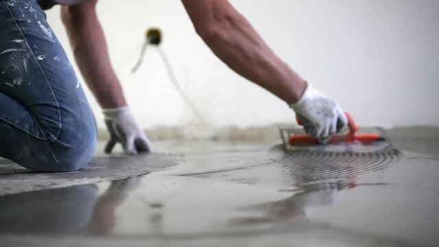 Building master in protective gloves pouring self-leveling screed on floor and spreading liquid cement solution with metal spatula. Repairman aligning cement mortar on construction site, finish of
