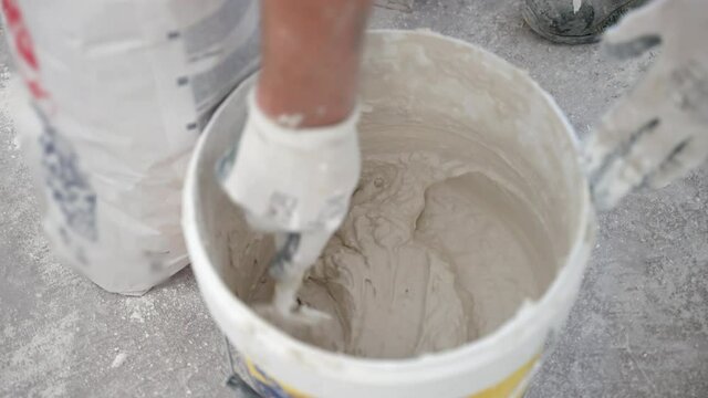 Workman in protective gloves using small metal spatula for mixing plaster solution in plastic bucket on construction site. Builder preparing mortar for putty walls of new apartment, mixture of cement