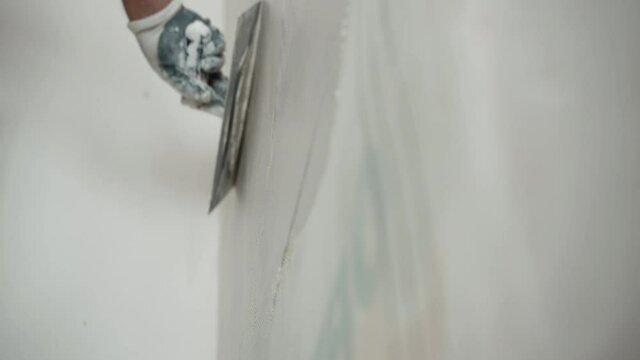 Repairing works on construction site, engineering worker in protective gloves spackling wall with metal spatula, man leveling and aligning wall surface with white finishing plastering paste solution