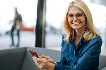 Smiling elegant middle aged caucasian woman in formal wear and eyeglasses uses smartphone, browsing internet or chatting with friends, looks at the camera, smiling