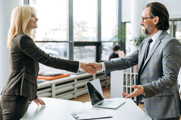 Satisfied business people are shaking hands after successful negotiations on briefing meeting. Influential colleagues in formal suits standing in modern office, coming to agreement