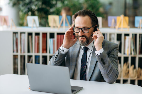 Smiling male operator of call center in headphones communicate with clients, consulting people. Senior business man sitting at the work desk, uses computr, having a conversation