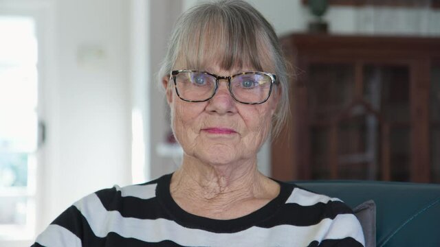 Close Up Portrait Of Senior Caucasian Woman At Home Looking At Camera. Older Woman With Grey Hair In Striped Sweater Sitting In Her Living Room. Slow Motion 4k