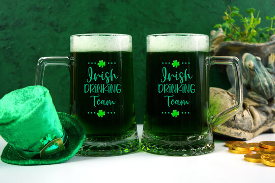 Happy St Patrick's Day two green beer steins, styled with leprechaun hat, shamrocks, and chocolate gold coins, against a textured green background. Drinking Team text on glass.
