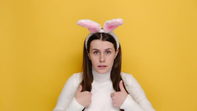Puzzled young female 20s in bunny fluffy ears, point fingers herself ask say who me no thanks i do not need it, wears white jumper, isolated on yellow background. Easter holiday and emotion concept