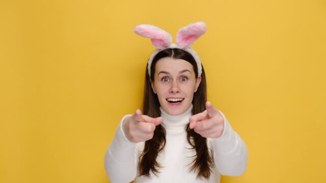 Side view of surprised woman 20s in bunny fluffy ears put hands on cheeks doing winner gesture showing thumb up, wears jumper, isolated on yellow studio background. Easter holiday and emotion concept