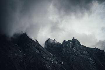 Dark atmospheric landscape with black rocky mountain wall in gray cloudy sky. Gray low cloud on top of black mountain. Dark mountain peak in low clouds in overcast weather. Gloomy minimalist landscape